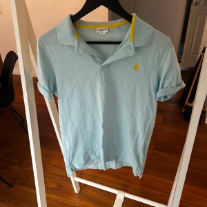 Ljusblå piké från U.S. Polo Assn. - Fräsch ljusblå pikétröja från U.S. Polo Assn. med gul broderad logga på bröstet och gul detalj på insidan av kragen. Klassisk krage och korta ärmar, perfekt för en chill och sportig look. Materialet är mjuk bomull som andas bra.