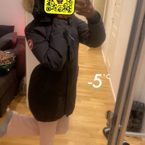 Svart dunjacka från Canada Goose - Säljer en svart lång dunjacka från Canada Goose med huva och snygg pälskant. Jackan har klassisk logga på ärmen, dragkedja och knappar framtill. Perfekt för kalla dagar och riktigt varm. Stilsäker och tidlös modell.