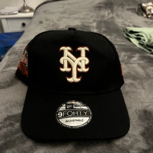 Svart New Era Mets keps 9FORTY - Säljer en svart New Era 9FORTY keps med broderad Mets-logga framtill i vitt och orange. Sidorna har broderade detaljer med Mets och årtal. Justerbar snapback baktill och böjd skärm med grön undersida. Perfekt för dig som gillar sportig streetstyle.