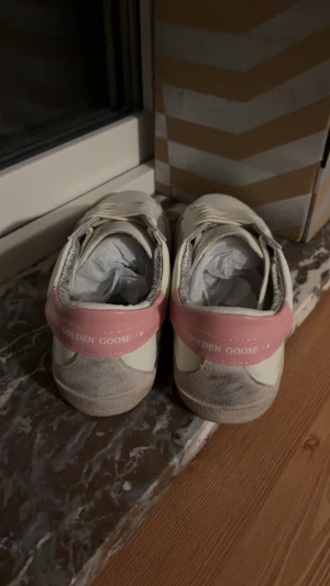 Golden Goose vita sneakers med rosa detalj - Golden Goose sneakers i vitt skinn med rosa hälparti och silverfärgad stjärna på sidan. Skorna har snörning, rund tå och platt sula. Insidan är fodrad med silverdetaljer och yttersulan är svart. Perfekt för dig som gillar coola och trendiga sneakers.