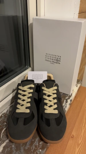 Maison Margiela svarta sneakers - Maison Margiela sneakers i svart skinn och mocka med beige snörning och gummisula. Klassisk rund tå och platt sula, med stilren design och neutrala färger. Perfekt för dig som gillar minimalistisk och trendig stil.