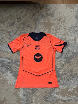 FC Barcelona third jersey, Pedri #8, Size: S/M - Säljer en officiell FC Barcelona matchtröja från Nike i orange med mörkblå detaljer. Tröjan har korta ärmar, V-ringad krage och tryck med Pedri #8 på ryggen. Materialet är lätt och andas, perfekt för fotboll. Spotify-logga på bröstet och UNHCR-tryck nedtill. New Condition, and player version  Size: S/M  ⚠️ Please send price proposal