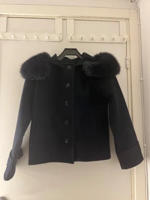 Mörkblå kappa med fuskpäls på huvan - Säljer en mörkblå kappa med fluffig fuskpäls på huvan och stora knappar framtill. Jackan har en rak och lite loose passform, långa ärmar och två fickor. Perfekt för dig som vill ha en stilren och mysig look under höst och vinter.