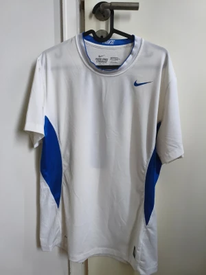 Nike Pro Combat vit/blå träningströja - Nike Pro Combat Dri-Fit t-shirt i vitt med blå detaljer och meshpaneler. Snygg fitted passform, korta ärmar och sportig look. Perfekt för träning med ventilerande material och coolt mönster på ryggen. Klassisk Nike-logga på bröstet. T-shirten är ny utan prislapp.