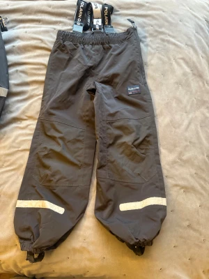 Svarta skalbyxor från Polarn O. Pyret - Svarta skalbyxor från Polarn O. Pyret med justerbara hängslen med logotyp, elastisk midja och reflexdetaljer vid bensluten. Byxorna har dragkedja i sidan och förstärkta knän, perfekta för kalla dagar utomhus.