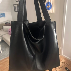 Svart axelväska i skinnimitation - Snygg svart axelväska med stilren design och breda handtag. Väskan har en praktisk dragkedja på sidan och en avtagbar axelrem. Materialet är skinnimitation som ger en cool och modern vibe. Perfekt storlek för att få plats med allt du behöver.