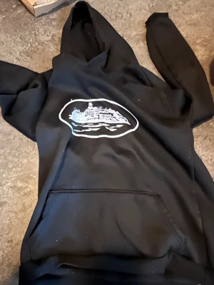 Svart cortez hoddie  - Svart hoodie med stor ficka framtill och tryck av ett fartyg i vitt och blått på bröstet. Tröjan har huva och långa ärmar, perfekt för dig som gillar coola motiv och avslappnad stil. Materialet känns mjukt och bekvämt mot huden.