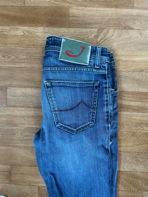 Jacob Cohen blå jeans style 622 - Snygga blå jeans från Jacob Cohen, modell style 622. Jeansen har en klassisk femficksdesign, röd broderad J på fickan och knappgylf. De är i stretchig denim med slitning och hål på vänster lår för en cool look.