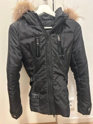 Svart pufferjacka med päls från Jayloucy - Svart pufferjacka från Jayloucy Paris med äkta päls på huvan, flera dragkedjor och snygga detaljer i skinnimitation. Jackan har figurnära passform, bältesdetaljer i midjan och långa ärmar med knappstängning. Perfekt för kalla dagar med stil.