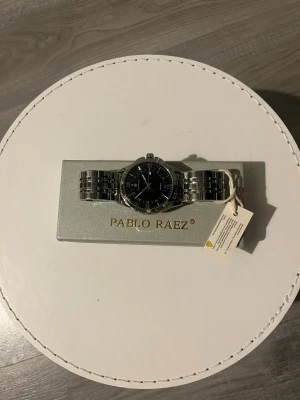Silverfärgad klocka Pablo Raez: helt oanvänd: med skyddsplast  - Stilren klocka från Pablo Raez med svart urtavla och romerska siffror. Armbandet är i blankt silverfärgat metall och har en klassisk länkdesign. Klockan har quartz-urverk och en rund form som ger en tidlös vibe. Perfekt för dig som gillar minimalistisk och modern stil.