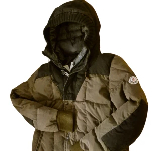 Moncler mörkgrön pufferjacka med huva - Säljer en fet pufferjacka från Moncler i mörkgrön och svart. Jackan har en stor huva med ribbad kant, quiltad design och dragkedja samt knappar framtill. Moncler-logga på ärmen och mjuka muddar vid handlederna. Perfekt för kalla dagar.