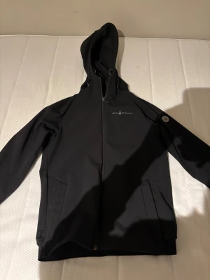 Svart vindjacka från Sail Racing - Svart vindjacka från Sail Racing med huva och hel dragkedja framtill. Jackan har diskret logga på bröstet och en rund detalj på ärmen. Snygg, stilren design med praktiska fickor och lång ärm. Perfekt för dig som gillar sportig och clean look.