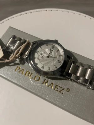 Helt ny oanvänd Pablo Raez Klocka! - Stilren klocka från Pablo Raez med rund urtavla i silver och romerska siffror. Armbandet är i blankt metallmaterial och har en klassisk länkdesign. Urtavlan har snygga linjer och datumvisning. Perfekt för dig som gillar en clean och modern look.