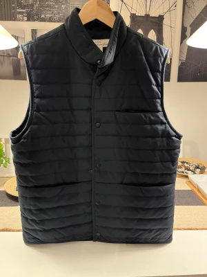 Mörkblå quiltad pufferväst från H&M L.O.G.G. - Mörkblå quiltad pufferväst från H&M L.O.G.G. i storlek M. Västen har en hög krage och horisontella sömmar som ger en snygg puffad look. Materialet är 100% polyester och insidan har ett meshfoder för extra komfort. Perfekt för lager-på-lager under höst och vinter. Helt ny utan prislapp.