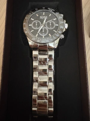 Silverfärgad  Hugo Boss klocka - Snygg klocka från Hugo Boss med svart urtavla och silverfärgad länkarmband i metall. Klockan har tre subdials, markerade index och tydliga siffror på bezeln. Boss-logga på både urtavla och spänne. Perfekt accessoar för dig som gillar stilren design.