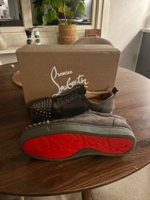 Christian Louboutin sneakers med nitar - Säljer ett par gråa sneakers från Christian Louboutin i mocka med ikoniska röda sulor och coola nitar på tån. Skorna har snörning och en rund tå, perfekt för dig som vill sticka ut med en edgy vibe. Kommer inte med originalkartong.