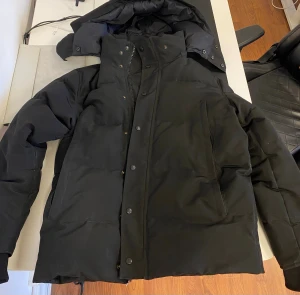 Svart Canada Goose jacka - Säljer min svarta  Canada Goose för att jag har vuxit ifrån den. Jackan har luva, dragkedja och tryckknappar framtill. Den är i bra kvalité och sällan använd 