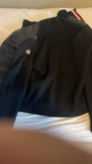 Svart pufferjacka från Moncler - Svart pufferjacka från Moncler med stickade partier och glansiga quiltade ärmar. Jackan har Moncler-logga på ärmen och en cool ribbad struktur på kroppen. Dragkedja framtill med röd, vit och blå detalj vid halsen. Perfekt för dig som gillar streetstyle.