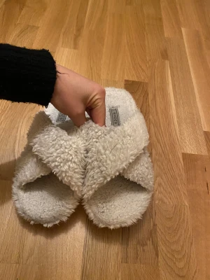 Fluffiga vita sandaler med korsad design - Supermysiga sandaler i fluffigt vitt material med korsad design över foten. Perfekta att glida runt i hemma för en chill vibe. Sulan är platt och hela skon är täckt av mjuk pälsimitation för extra komfort.