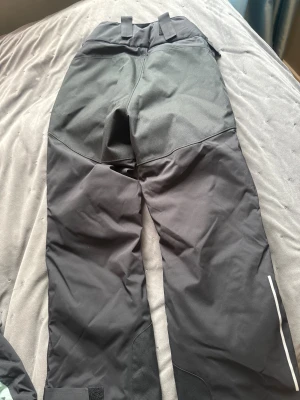 Svarta skidbyxor med reflexdetalj - Svarta täckbyxor med förstärkta partier på knän och bak, justerbara kardborreband vid benslut och midja, samt dragkedjor vid fickorna. Materialet är slitstarkt och vattenavvisande, perfekt för kalla dagar. Snygg reflexdetalj på benet för extra synlighet.