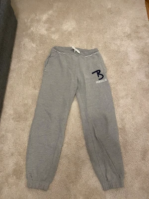 Grå mjukisbyxor Bondelid strl 160 - Snygga grå mjukisbyxor från Bondelid med vit snörning i midjan och elastiska muddar vid bensluten. Byxorna har fickor och en stor mörkblå Bondelid-logga på ena benet. Perfekta för chill och häng med kompisar.