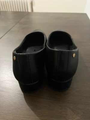 Svarta ballerinaskor med låg klack - Stilrena svarta ballerinaskor från So All med spetsig tå och låg klack. Skorna är tillverkade i skinnimitation och har en elegant, minimalistisk design med en liten guldfärgad detalj bak på hälen. Perfekt för dig som gillar klassisk och enkel stil.