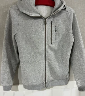 Grå hoodie från Sail Racing med dragkedja - Snygg grå hoodie från Sail Racing med hel dragkedja och huva med snörning. Tröjan har en bröstficka med dragkedja och logga på framsidan. Insidan är mjuk och fleecefodrad för extra komfort. Perfekt för chill dagar och sköna promenader. Använd några gånger. 