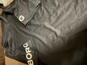 Svart Björn Borg t-shirt Hydro-Pro - Svart t-shirt från Björn Borg med Hydro-Pro teknologi. Snyggt tryck med BORG i vitt på ena ärmen och ett G på den andra. Klassisk rund hals och korta ärmar, perfekt för dig som gillar sportig stil och vill ha något bekvämt.