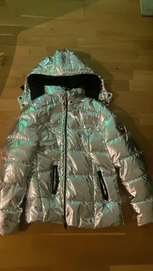 Silvrig pufferjacka med huva - Säljer en silvrig pufferjacka med huva och svarta detaljer vid fickorna. Jackan har quiltad design, dragkedja framtill och ett märke på ärmen. Perfekt för dig som vill sticka ut i vinter och hålla dig varm. Jackan är aldrig använd!