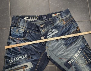 Japrag JP-111D jeans med tryck och nitar - Unika mörkblå jeans från Japrag JP-111D med vita tryck, kontrastsömmar, nitar och flera dragkedjor. Snygga detaljer på fickorna, broderade mönster och coola texttryck. Passar dig som vill sticka ut med en streetwear-look. Materialet är klassisk denim.  Mått: Midjemått: 43 cm (mätt platt) Höft: ca 56 cm Innerbenslängd: 84 cm Bredd vid låret: 30 cm Skrev: 24 cm Stuss: 37 cm Ytterbenslängd: 113 cm Benvidd längst ner: 20 cm  Skriv för frågor, pris kan diskuteras ❤️  
