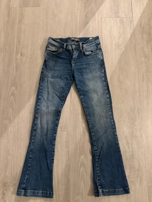 Blå bootcut jeans från LTB - Snygga blå jeans från LTB med bootcut passform och klassisk femficksdesign. Jeansen har en lätt tvättad look med slitningar och kontrastsömmar, samt detaljerade bakfickor. Perfekt för dig som gillar retrovibbar och bekväm stil.