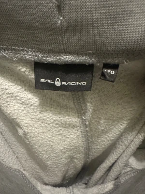 Grå mjukisbyxor från Sail Racing - Snygga grå mjukisbyxor från Sail Racing med logga längs benet. Byxorna har ribbad midja med snörning, en bakficka. Perfekt för chill eller träning. Passformen är normal och benen är raka med ribbade muddar vid anklarna.