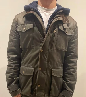 Mörkgrön Field jacket från Bondelid - Snygg mörkgrön field jacket från Bondelid med quiltat foder, flera stora fickor med knappar och dragkedja framtill. Jackan har en krage i manchester och detaljer i brunt. Perfekt för dig som gillar klassisk stil med moderna inslag. Jackan har även slitningar runt sig som ger dens snygga wash 