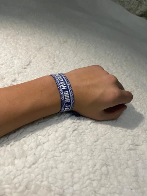 Blått armband Christian Dior J'Adior - Snyggt blått tygarmband från Christian Dior med texten 'J'Adior' broderad i vitt. Armbandet har justerbar knytning med tofsar i ändarna och en cool, sportig känsla. Perfekt accessoar för att lyfta din outfit med en touch av lyx.