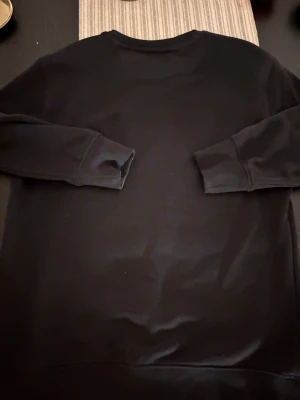 Svart sweatshirt från GANT, S - Svart sweatshirt från GANT i storlek S. Tröjan har rund hals med ribbad kant och en diskret broderad logga på bröstet. Insidan är mjuk och materialet känns som bomull. Kragen har snygga ränder i rött, vitt och blått för extra detalj.