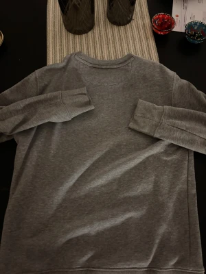 Grå sweatshirt från GANT, storlek S - Snygg grå sweatshirt från GANT i storlek S. Tröjan har rund hals, ribbade muddar och ett litet broderat GANT-emblem på bröstet. Tillverkad i mjuk bomull, perfekt för chill dagar. Enkel och stilren design som funkar till jeans eller joggers.