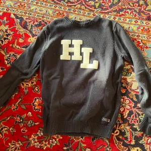 Stickad tröja från Henri Lloyd - Marinblå stickad tröja från Henri Lloyd med stora vita HL-bokstäver på bröstet. Rund halsringning med ribbad kant och långa ärmar. 