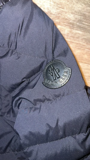 Svart Moncler pufferjacka herr - Svart pufferjacka från Moncler med quiltade partier på axlar och bröst, samt snygga silverfärgade dragkedjor på fickorna. Jackan har ståkrage och Moncler-logga på ärmen. Materialet är polyester och dun, perfekt för kyliga dagar.