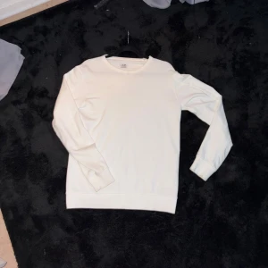 sweatshirt cp Company - Säljer en stilren vit sweatshirt från C.P. Company med deras ikoniska svart linsdetalj på ärmen. Tröjan har rund hals, långa ärmar och är gjord i mjuk bomull. Perfekt för dig som gillar cleana och moderna plagg med cool branding.självklart äkta