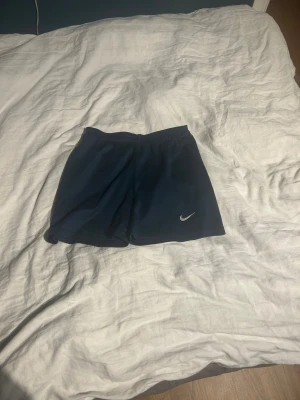 Mörkblå Nike shorts strl 16 - Säljer ett par mörkblå sportiga shorts från Nike med elastisk midja och klassisk vit swoosh-logga på benet. Perfekta för träning eller chill dagar. Lätta och bekväma med enkel design.
