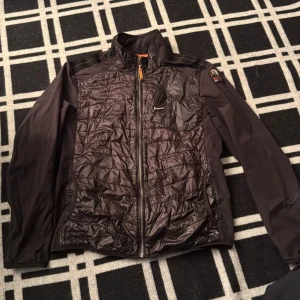 Svart quiltad jacka Parajumpers L - Svart jacka från Parajumpers i storlek L med quiltad framsida och rygg, samt släta ärmar. Jackan har dragkedja framtill, en bröstficka med zip och en liten patch på axeln. Materialet är mestadels polyester med lite elastan för extra komfort.