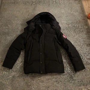 Svart dunjacka från Canada Goose - Svart dunjacka från Canada Goose med huva och klassisk logga på ärmen. Jackan har långa ärmar, flera fickor med dragkedja och tryckknappar framtill. Perfekt för kalla dagar och har en clean, stilren look som funkar till allt.