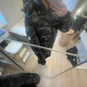 Svart Moncler pufferjacka med huva - Säljer en svart, glansig pufferjacka från Moncler med huva och dragkedja framtill. Jackan har lång ärm, quiltad design och klassisk Moncler-logga på ärmen. Perfekt för kalla dagar och har en riktigt snygg streetstil.