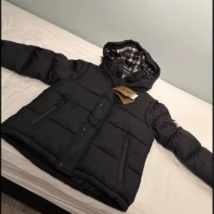 Svart pufferjacka från Burberry - Svart pufferjacka från Burberry med stor huva fodrad i rutigt tyg i grått, vitt och rött. Jackan har dragkedja och knappar framtill, två sidofickor med dragkedja och tydlig Burberry-logga på ärmen. Perfekt för kalla dagar med sin tjocka och quiltade design.