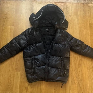 Svart pufferjacka Alessandro Zavetti - Svart pufferjacka från Alessandro Zavetti med glansig finish och stor huva. Jackan har dragkedja framtill, två fickor med dragkedja och snyggt mönstrat innerfoder med logotyp. Perfekt för kalla dagar och har en modern, urban vibe.