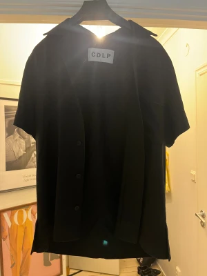  CDLP - Svart skjorta från CDLP med klassisk krage och korta ärmar. Skjortan har knappar framtill och en stilren, loose passform som ger en chill vibe. Perfekt för dig som gillar minimalistisk stil och vill ha något enkelt men snyggt.