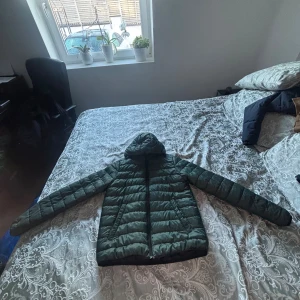 Grön pufferjacka med huva - Säljer en mörkgrön pufferjacka med huva och dragkedja framtill. Jackan har ett quiltat mönster och långa ärmar, perfekt för kyliga dagar. Enkel och stilren design som funkar till många olika outfits.