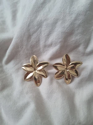 Guldiga blomformade örhängen - Säljer ett par stora, guldiga örhängen i form av blommor med sex blad. De har en blank finish och fästs med klassisk stift. Perfekta för dig som vill ha ett statement-smycke med cool blomdesign.