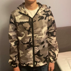 Camouflage vindjacka från Lindfors - Säljer en snygg vindjacka från Lindfors med grå, beige och svart camouflage-mönster. Jackan har huva med snörning, hel dragkedja framtill och elastiska muddar vid ärmslut. Perfekt för dig som gillar streetstyle och vill sticka ut.