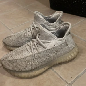 Adidas Yeezy Boost 350 V2 ljusgrå - Säljer ett par Adidas Yeezy Boost 350 V2 sneakers i ljusgrå färg med stickad ovandel och genomskinlig, ribbad platt sula. Skorna har snörning och en rund tå, samt den klassiska boost-dämpningen för extra komfort. Perfekt för dig som gillar streetwear och vill sticka ut.